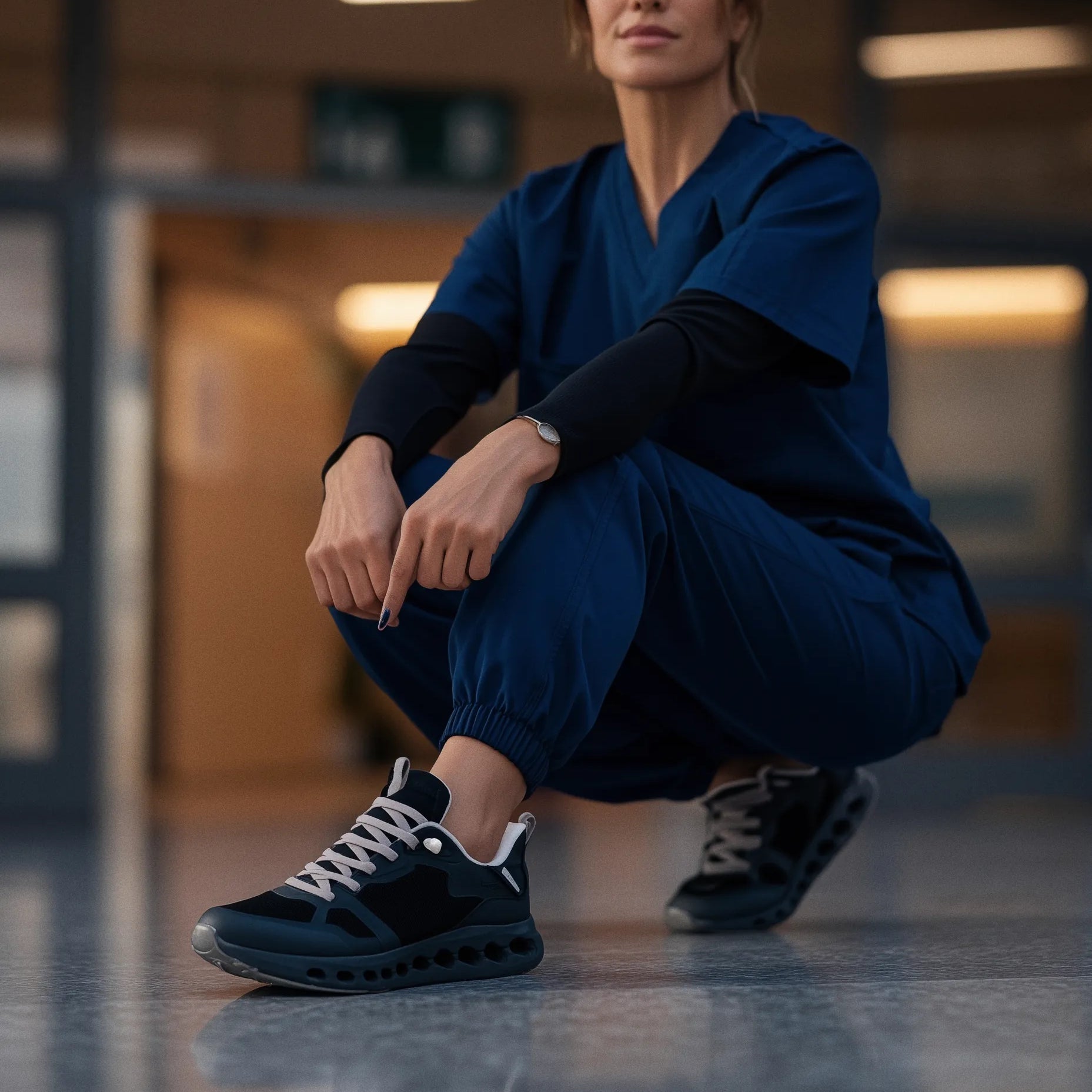 A woman in navy blue medical scrubs crouches in a hallway, wearing black and navy athletic sneakers with white laces.
