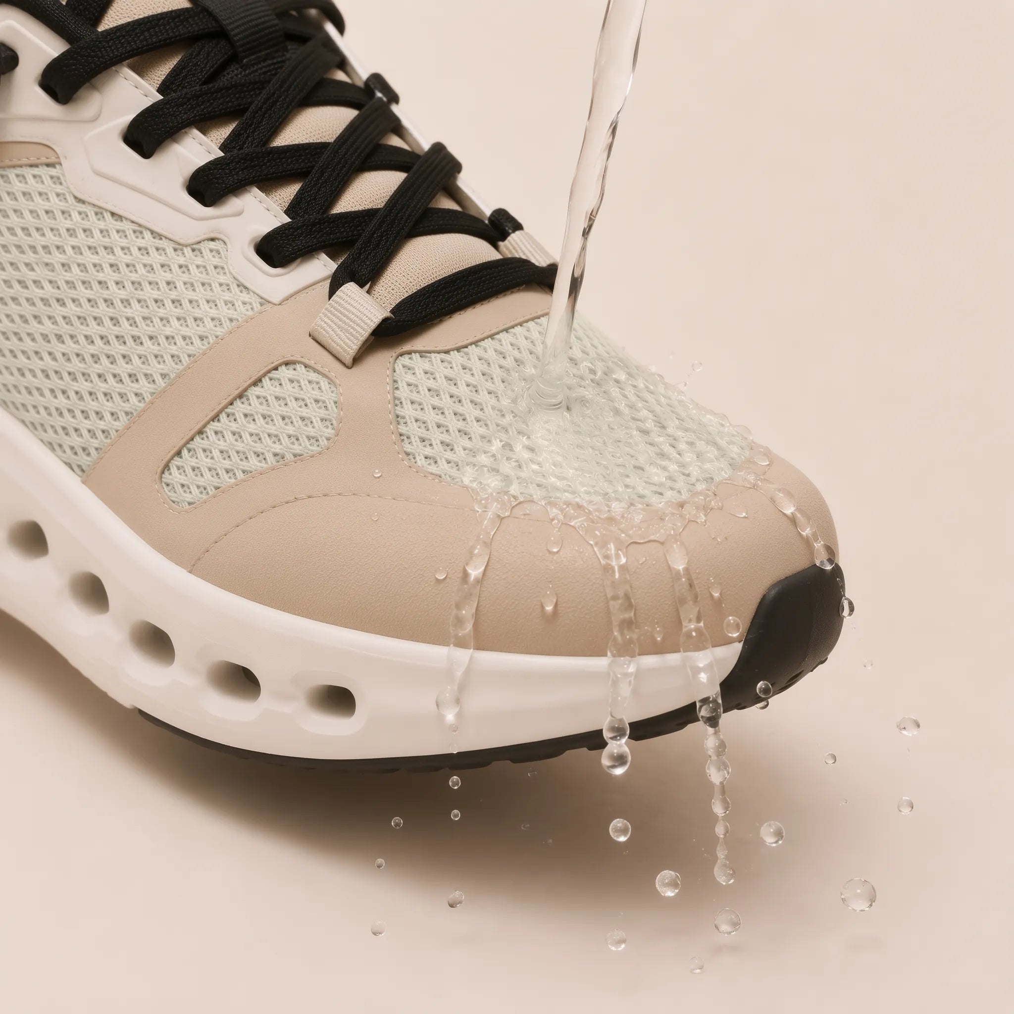 A close-up of a sneaker with water being poured over it, showing water droplets sliding off its textured upper and midsole. The shoe features a light beige and mint green mesh upper with black laces and a white cushioned sole.