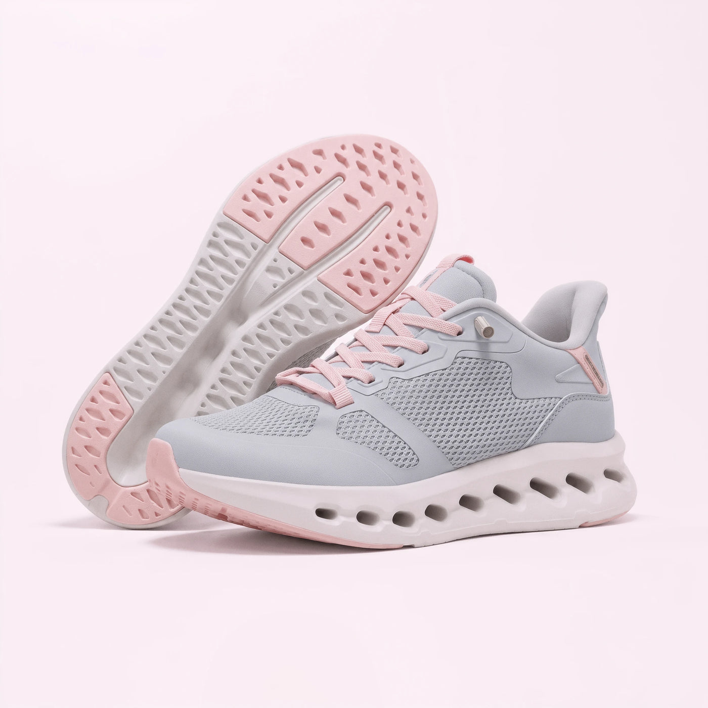 A pair of light gray and pink athletic sneakers, with one shoe standing upright and the other lying on its side to show the sole, set against a solid pale pink background.
