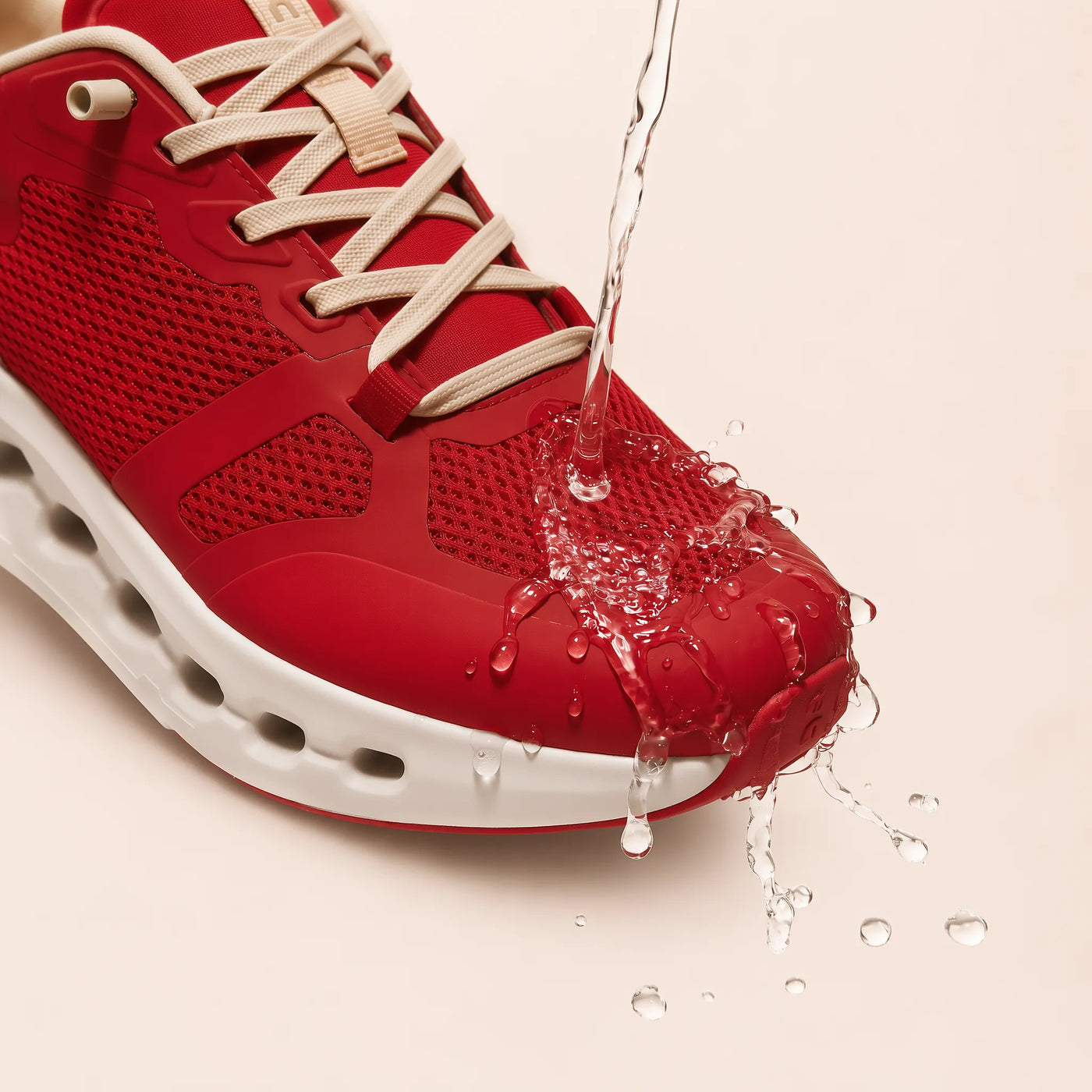 A close-up of a red athletic shoe with beige laces and a white sole, shown with water pouring over the toe area to demonstrate water resistance, on a light beige background.