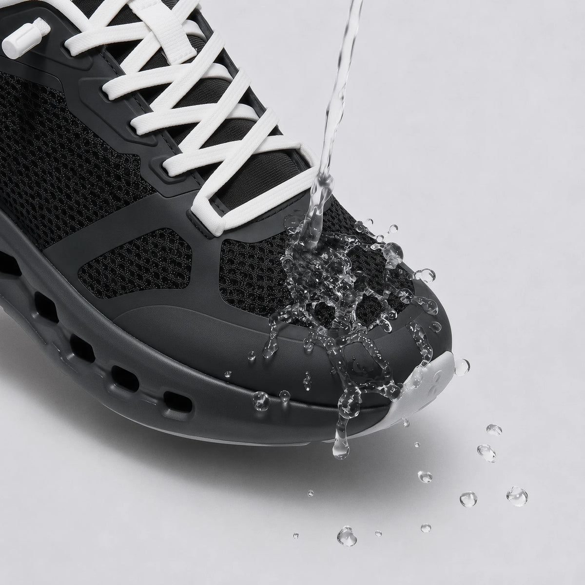 A close-up of a black athletic sneaker with white laces, shown with water being poured over the toe area to demonstrate water resistance, on a light gray background.