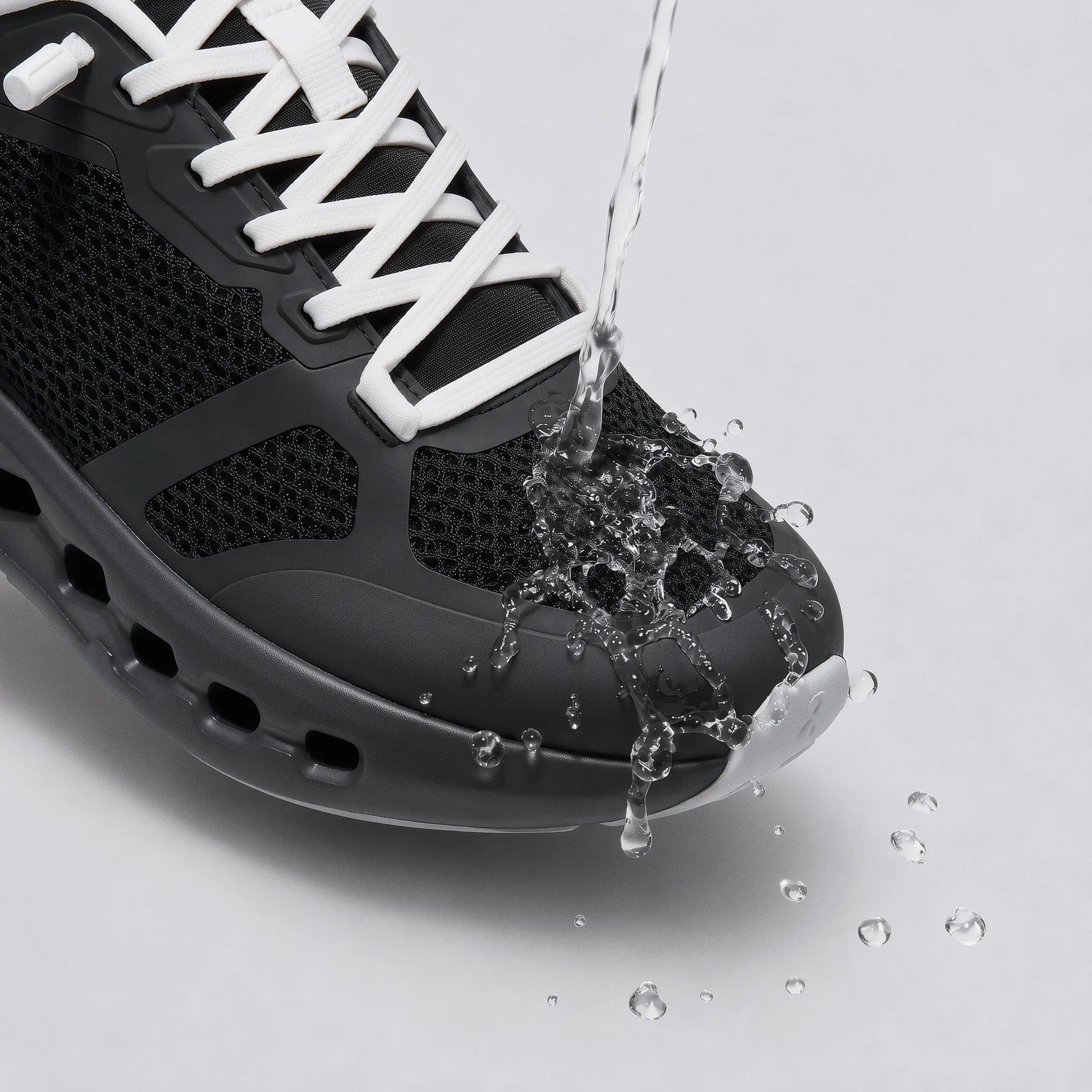 A close-up of a black athletic sneaker with white laces, shown with water being poured over the toe area to demonstrate water resistance, on a light gray background.