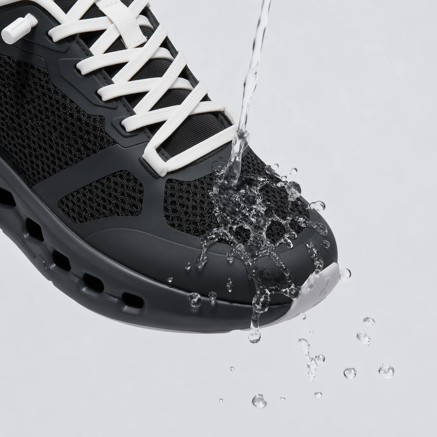 A close-up of a black athletic shoe with white laces, shown with water splashing over the toe area on a light gray background.