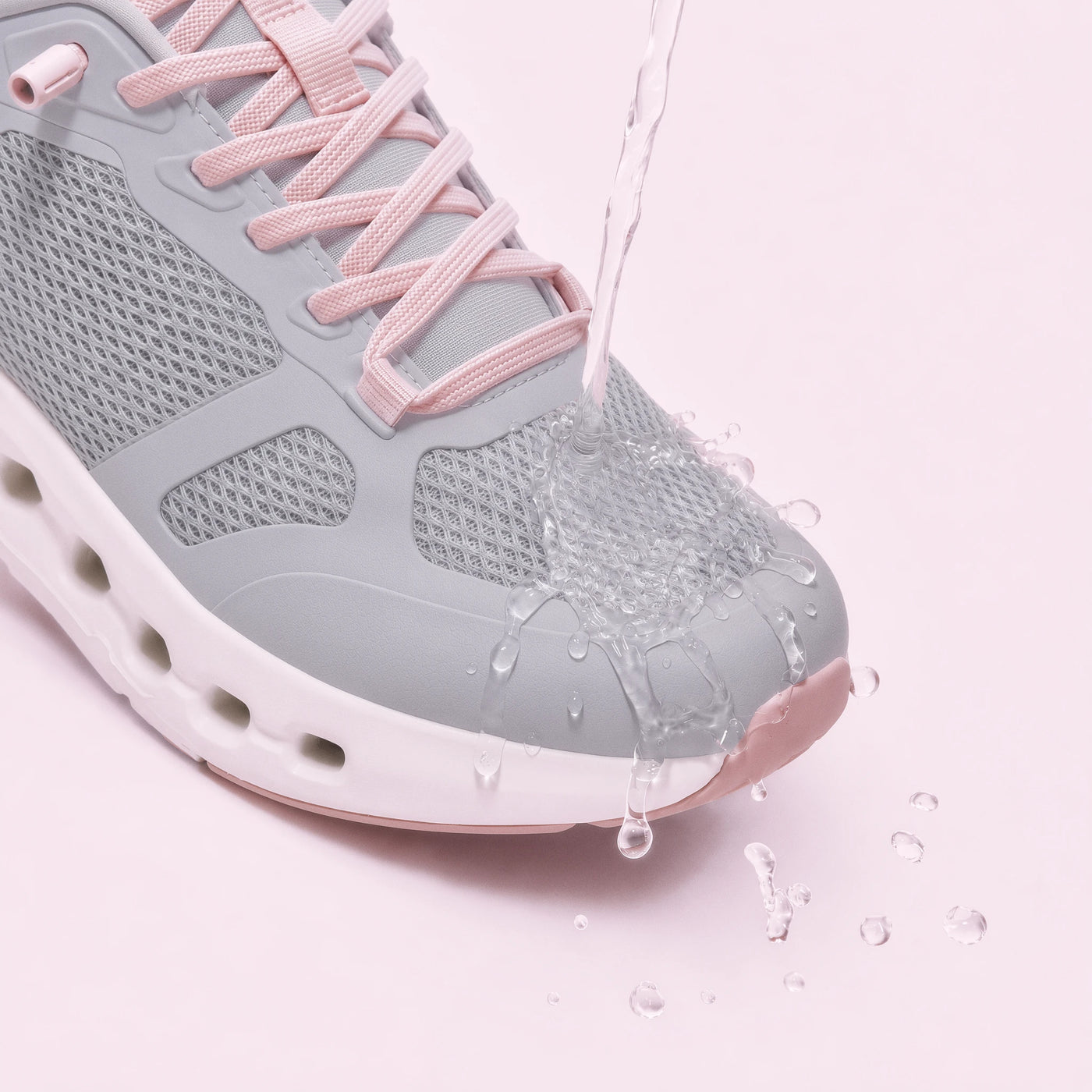 A close-up of a light gray and pink athletic sneaker with water being poured over the toe area to demonstrate water resistance, set against a solid pale pink background.