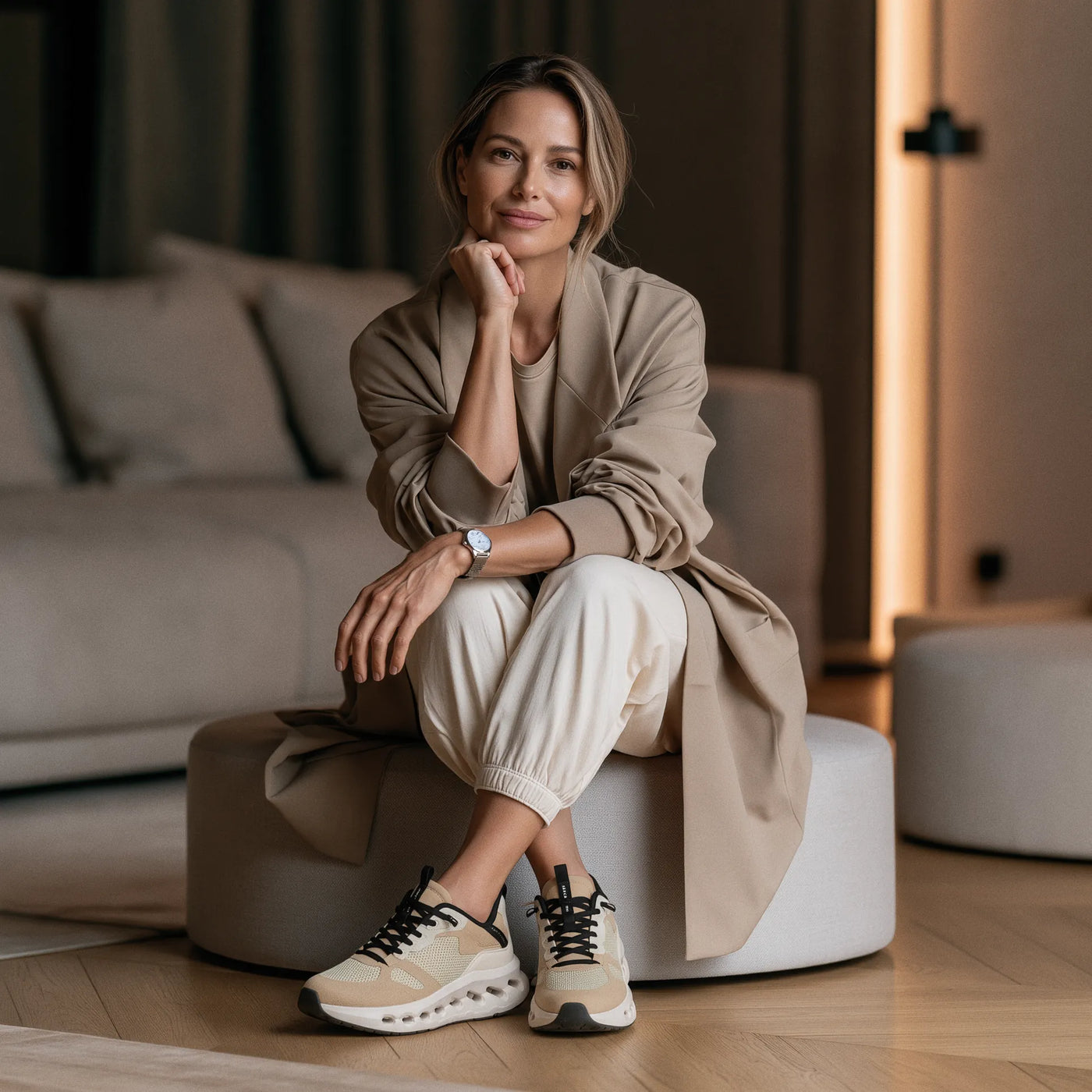 A woman sits on a round ottoman in a modern living room, wearing a beige trench coat, cream pants, and beige-and-black sneakers.